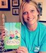 Smiling blonde with green shirt holding copy of her published book in her home